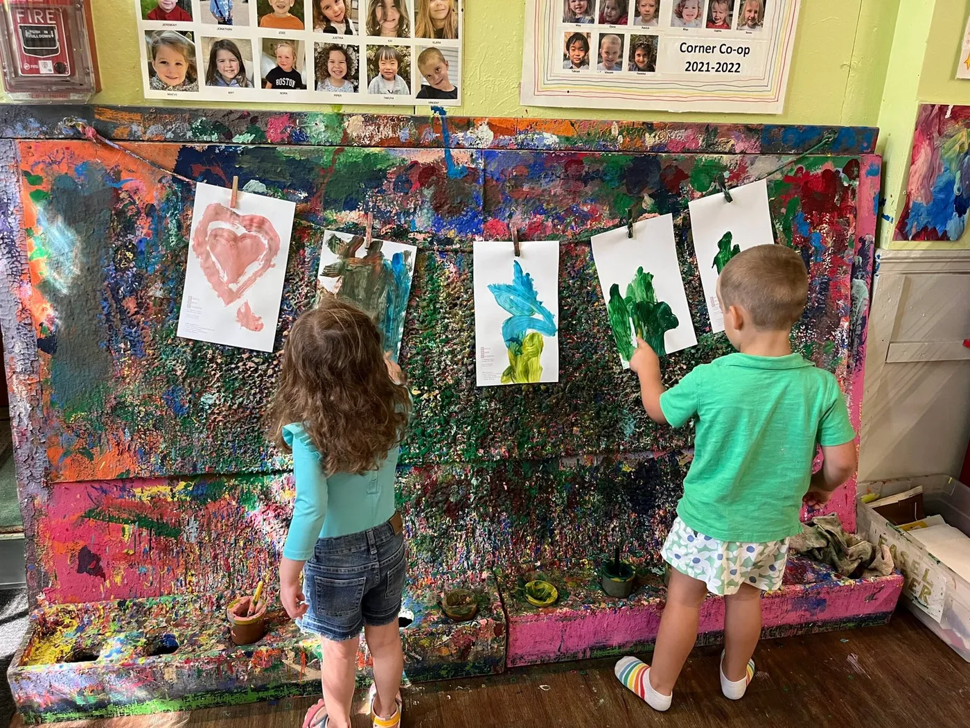 Two children painting at a large paint-splattered easel wall at Corner Co-op Nursery School