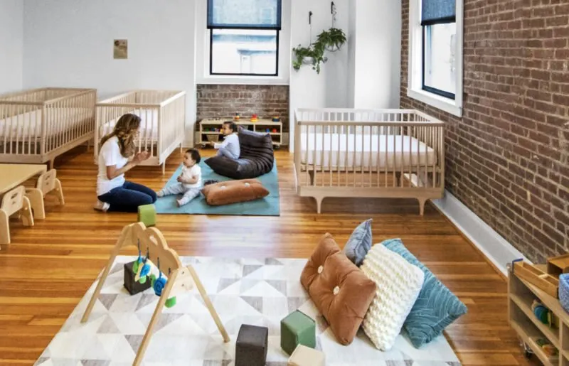 Infant room with cribs, floor mats and a teacher with a baby at Ethos in South Boston