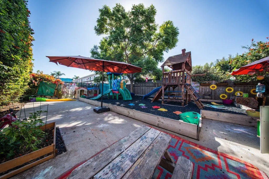 Shaded play yard with a wooden climbing structure and garden beds at Green Beans Preschool